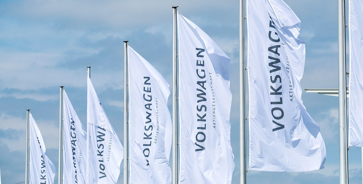 Es sind sieben Fahnenmästen zu sehen. Die weißen Fahnen tragen die dunkelblaue Aufschrift "Volkswagen - Aktiengesellschaft". Die Fahnen ragen in den hellblauen, leicht bewölkten Himmel.