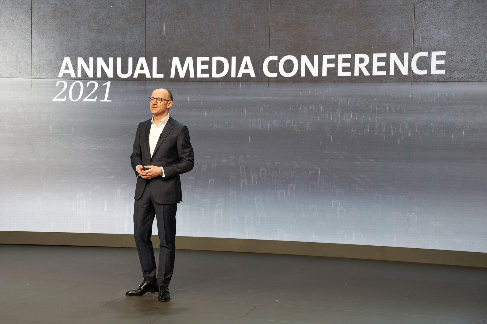 A man with glasses and a receding hairline stands on stage at the "Annual Media Conference 2021". He wears a dark suit with a white shirt and a clip-on microphone. The background features a large screen displaying the conference title and a modern graphic design.