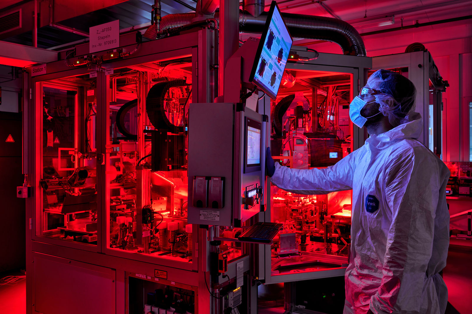 A technician in protective clothing monitors a red-lit high-tech manufacturing machine via a display.