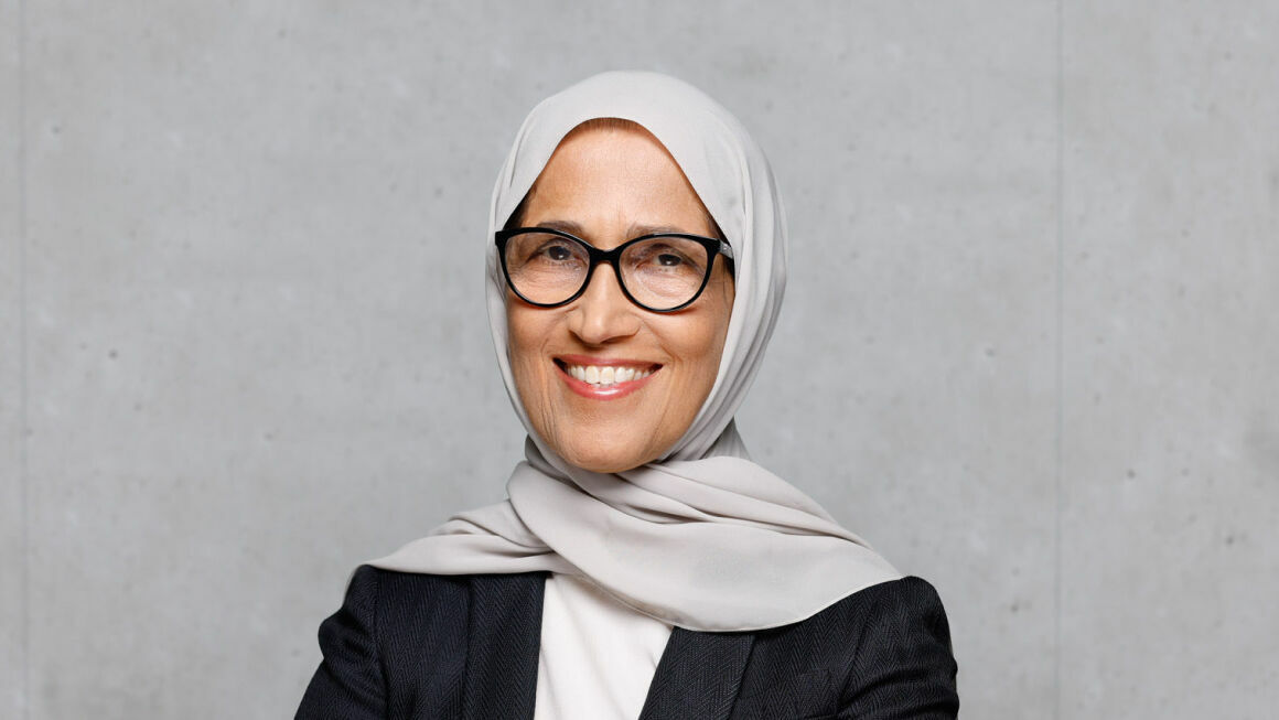 Eine lächelnde Frau mit beiger Hijab und schwarzer Brille steht vor einer grauen Betonwand. Sie trägt einen schwarzen Blazer über einem weißen Oberteil und hat die Arme entspannt verschränkt. Ihr freundlicher Gesichtsausdruck strahlt Professionalität und Selbstbewusstsein aus.
