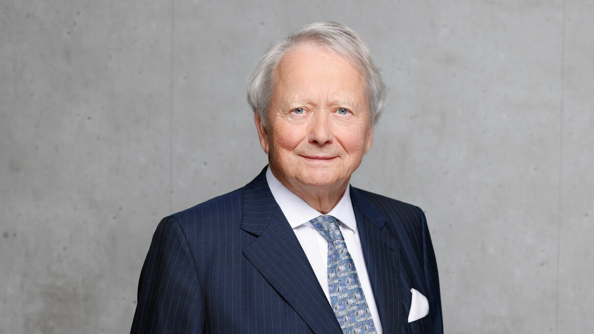 Das Bild zeigt Dr. rer. comm. Wolfgang Porsche vor einer grauen Beton-Wand. Er trägt einen Anzug, ein Hemd und eine gemusterte Krawatte.