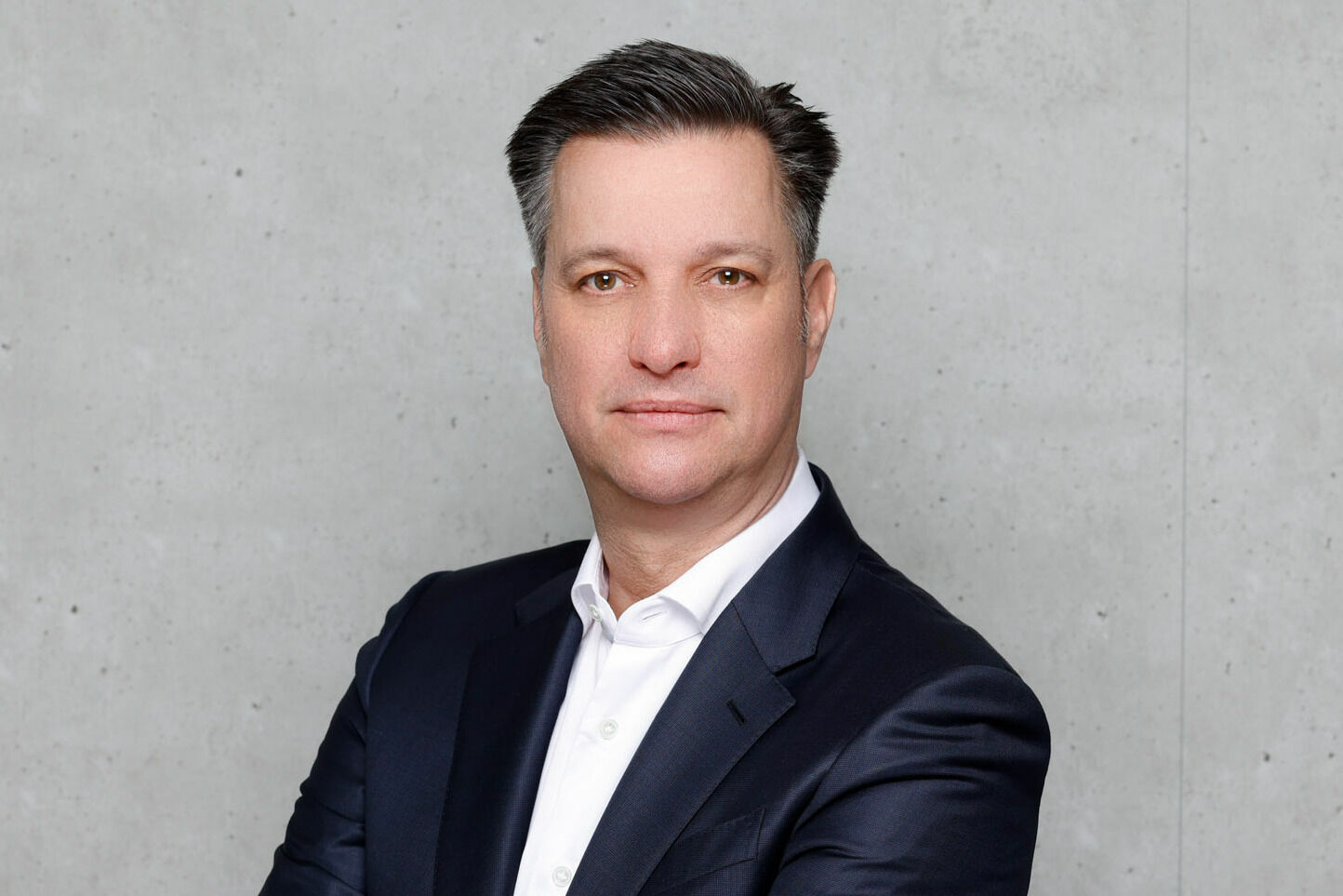 Das Bild zeigt Thomas Schmall-von Westerholt vor einer grauen Beton-Wand. Er trägt einen Anzug und ein Hemd.