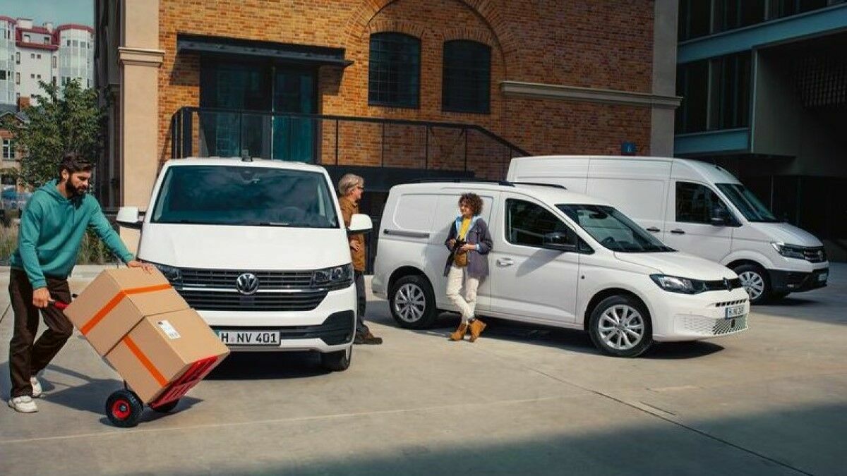 Three white Volkswagen commercial vehicles, including a Transporter, a Caddy, and a Crafter, are parked in an urban courtyard with a brick building. A man is moving packages with a hand truck, while two other people are engaged in conversation.