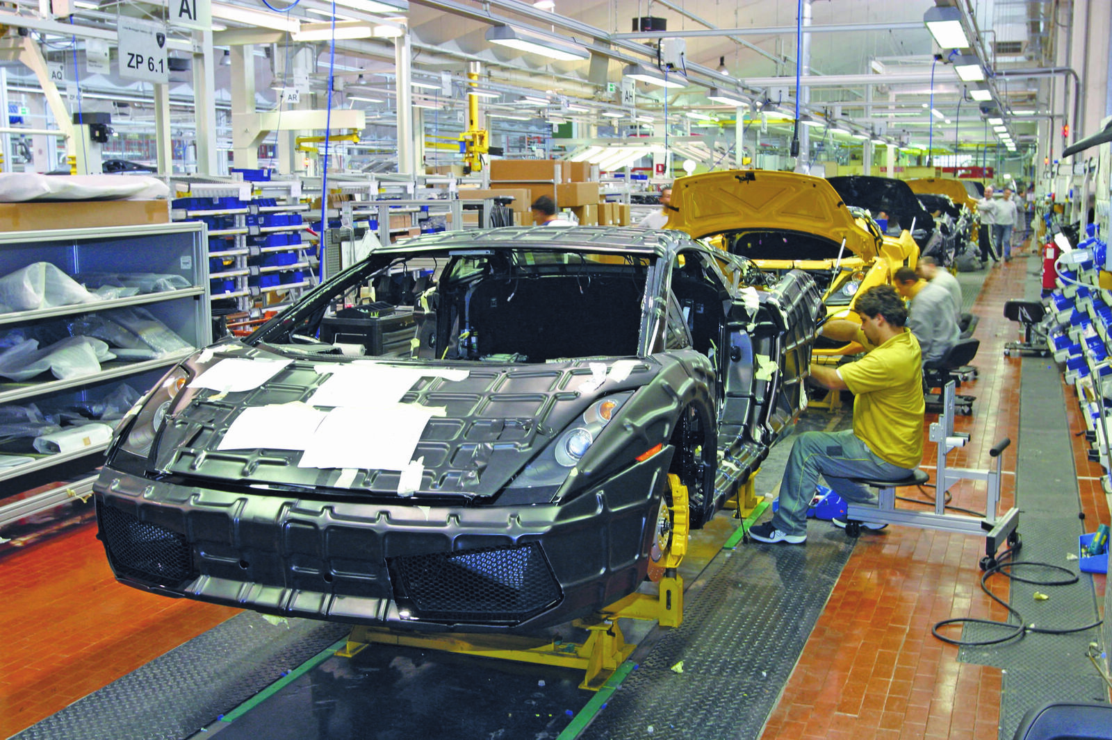 Assembly of a sports car in a modern automotive factory. Workers in yellow shirts are installing components on the body of a black supercar, while additional vehicles in various stages of production are visible along the assembly line.