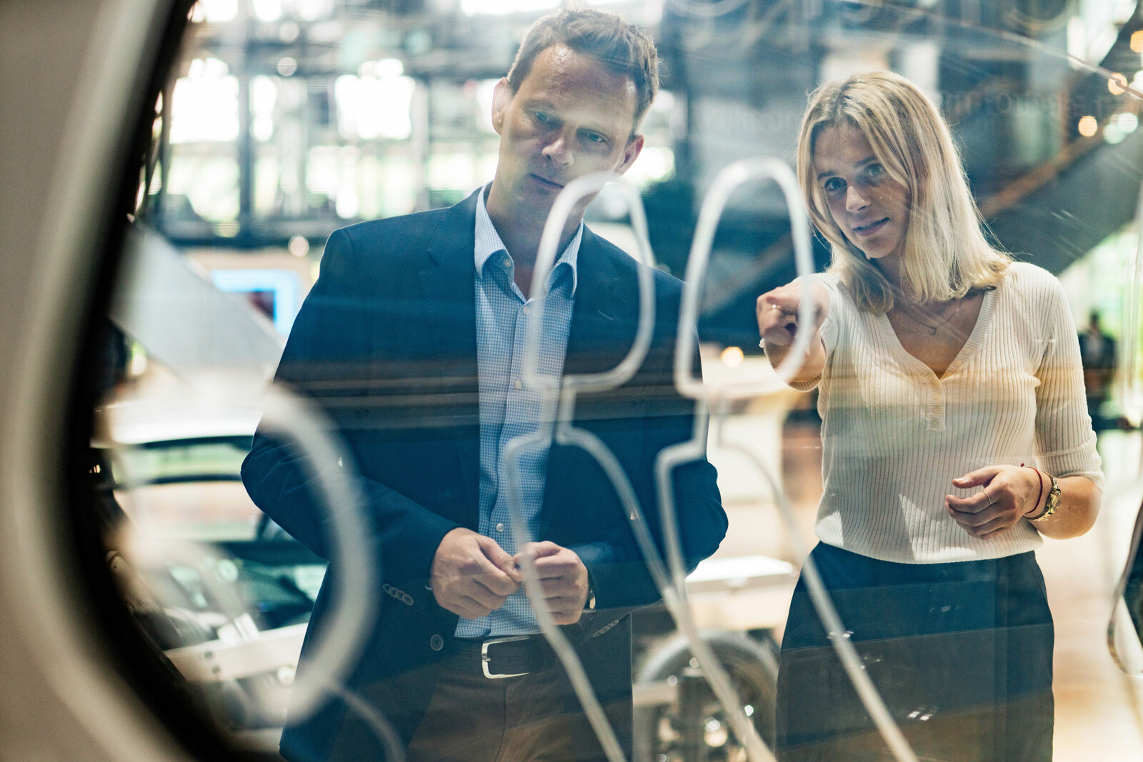 A man in a suit and a woman in a white top stand in front of a glass panel with sketched lines, engaged in discussion. The reflection reveals a modern environment with cars and large windows.