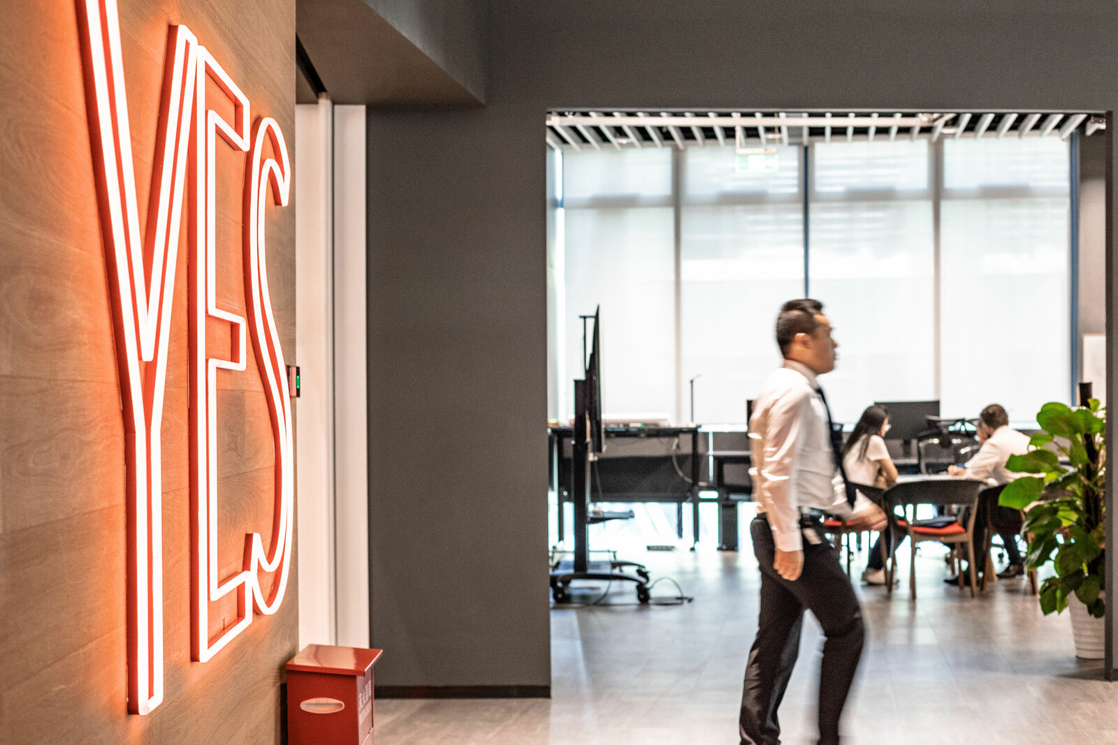 A modern office with an open design. In the foreground, a large red neon sign with the word 'YES' glows on the wall. A man in business attire walks through the space, while in the background, several people are working at a meeting table.