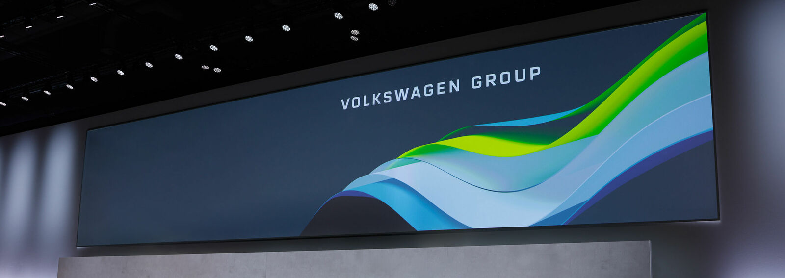 Large digital screen displaying the text “VOLKSWAGEN GROUP.” Below the text, a graphic features flowing, colorful lines in green, blue, and white on a dark background.