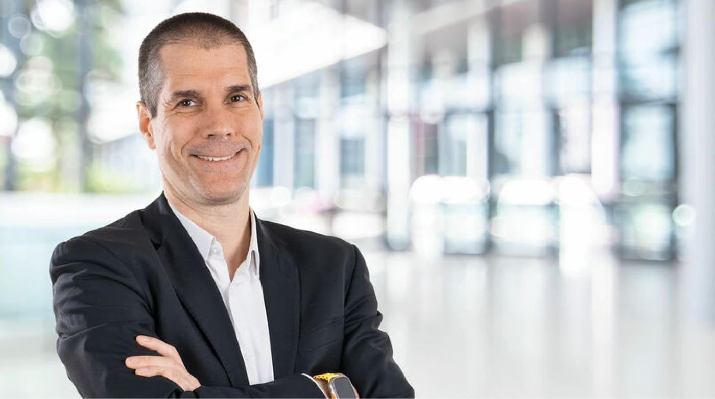 Portrait of a smiling man in business attire with arms crossed in front of a modern, light-filled office building.