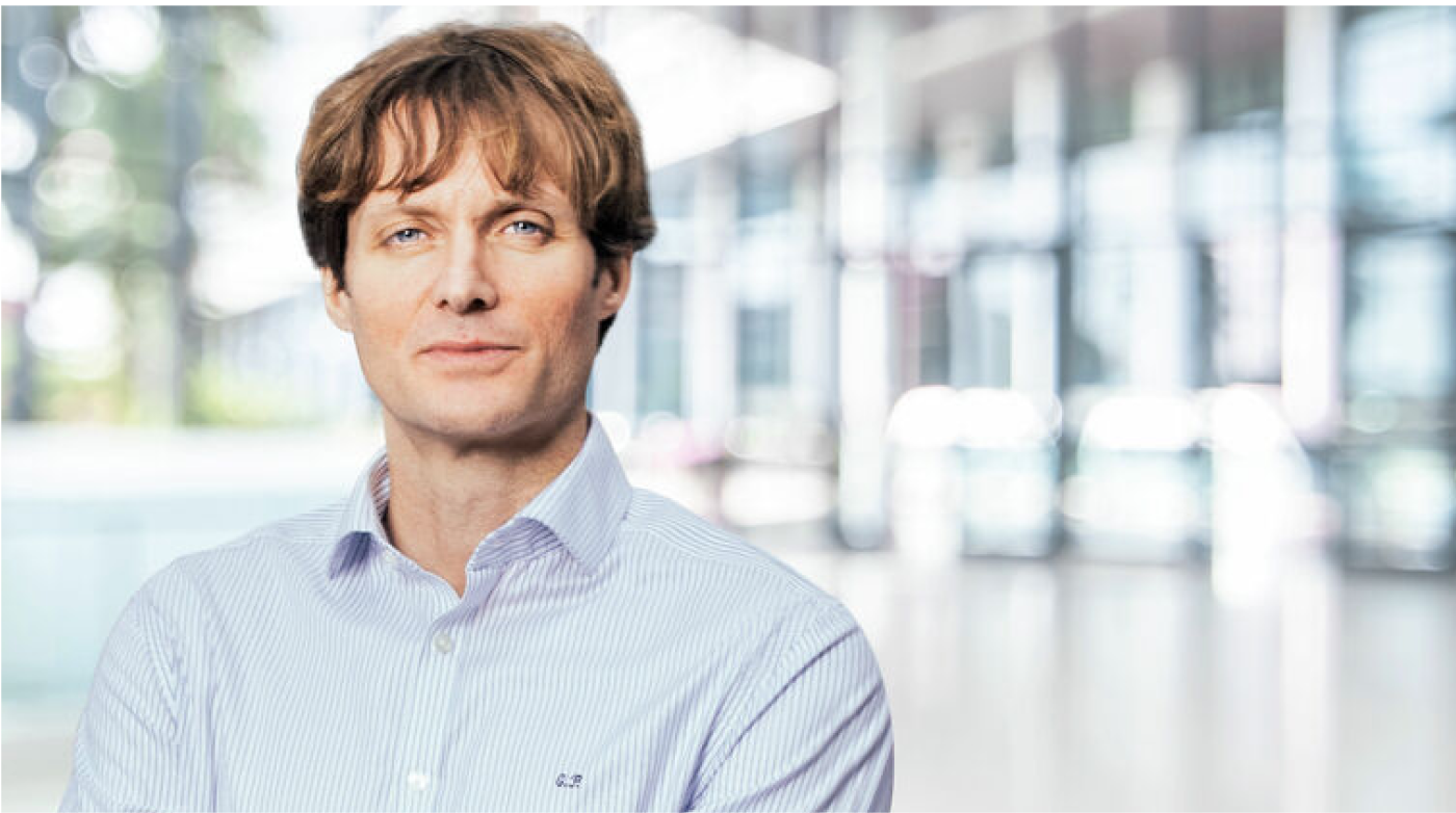 A man with medium-length, wavy hair wears a light blue striped shirt and looks at the camera with a friendly expression. The background features a modern, light-filled office environment.