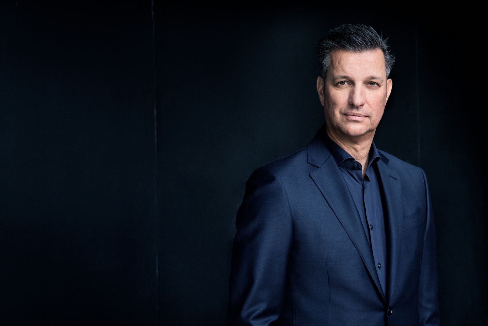 The picture shows Thomas Schmall in front of a black background. He is wearing a dark suit and a dark shirt.