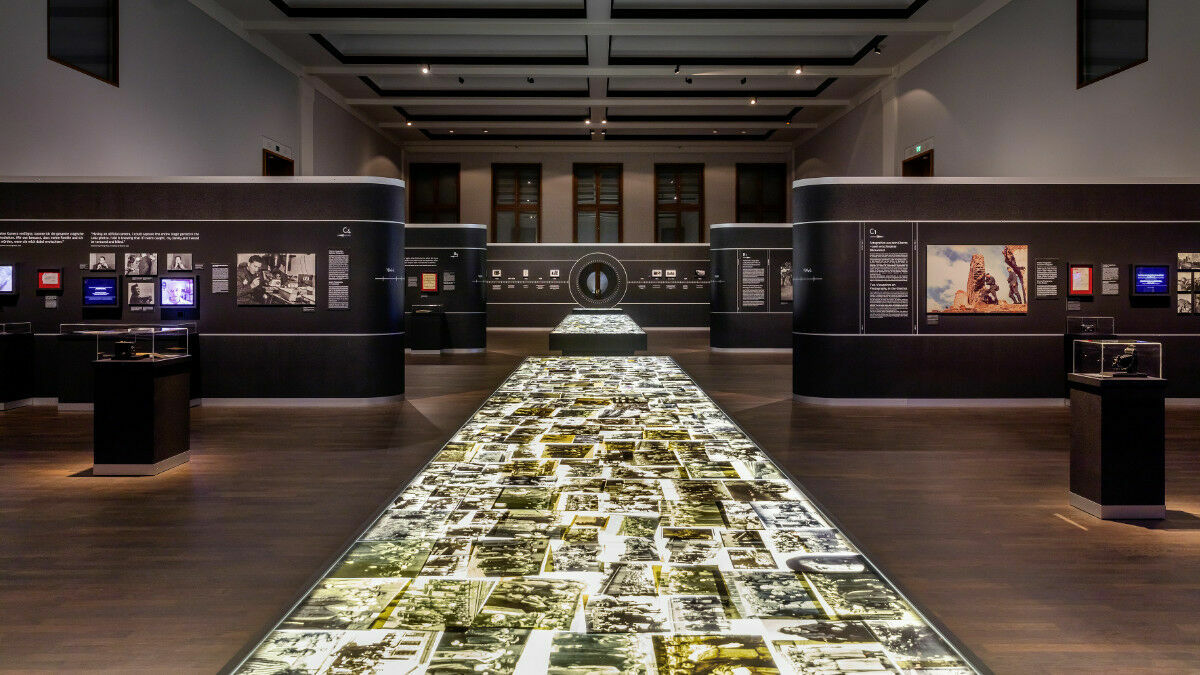 Interior view of a modern exhibition featuring an illuminated floor covered with historical photographs. The dark walls display informational panels, screens, and framed images.