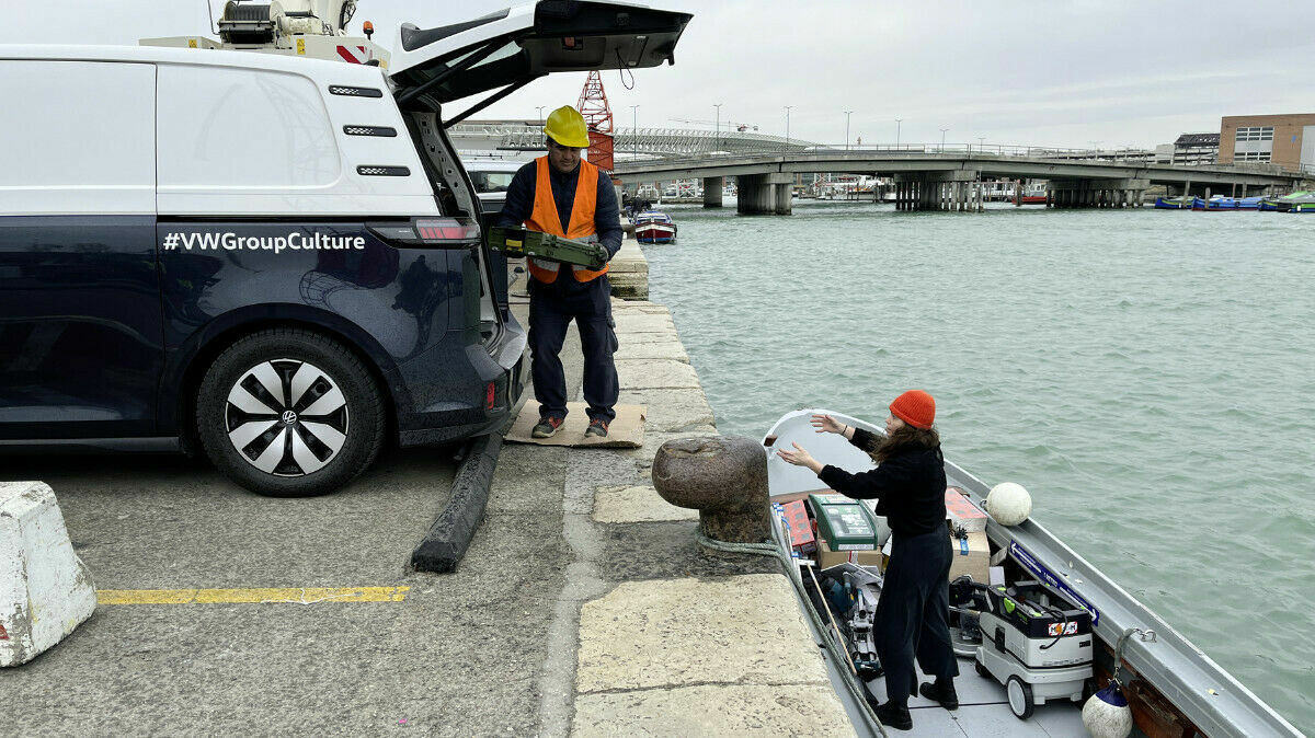 A man wearing a safety vest and a yellow hard hat stands by the open trunk of a VW van labeled "#VWGroupCulture," handing a box to a woman on a boat. The woman is wearing a red hat and dark clothing. In the background, a bridge and several boats on the water are visible.