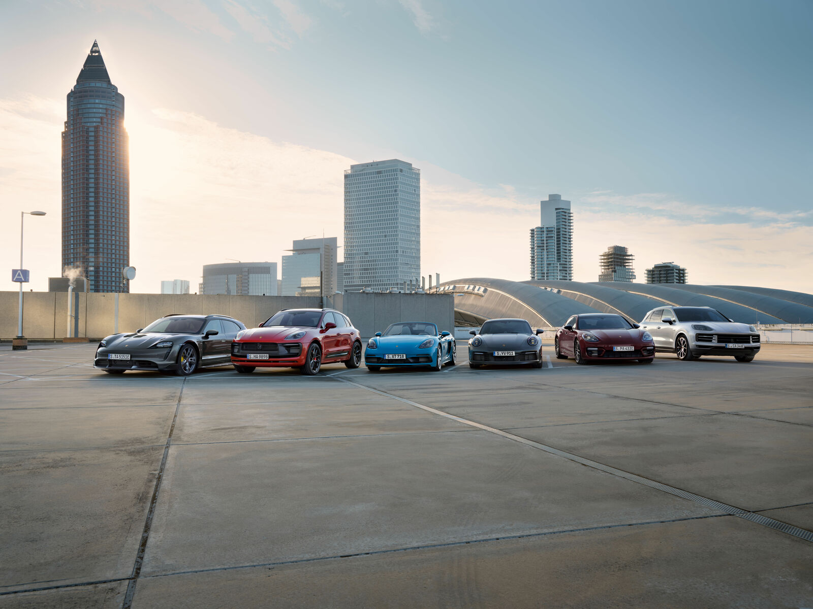Six Porsche models in different colors are parked side by side on a rooftop parking lot with a modern skyline and high-rise buildings in the background at sunset.