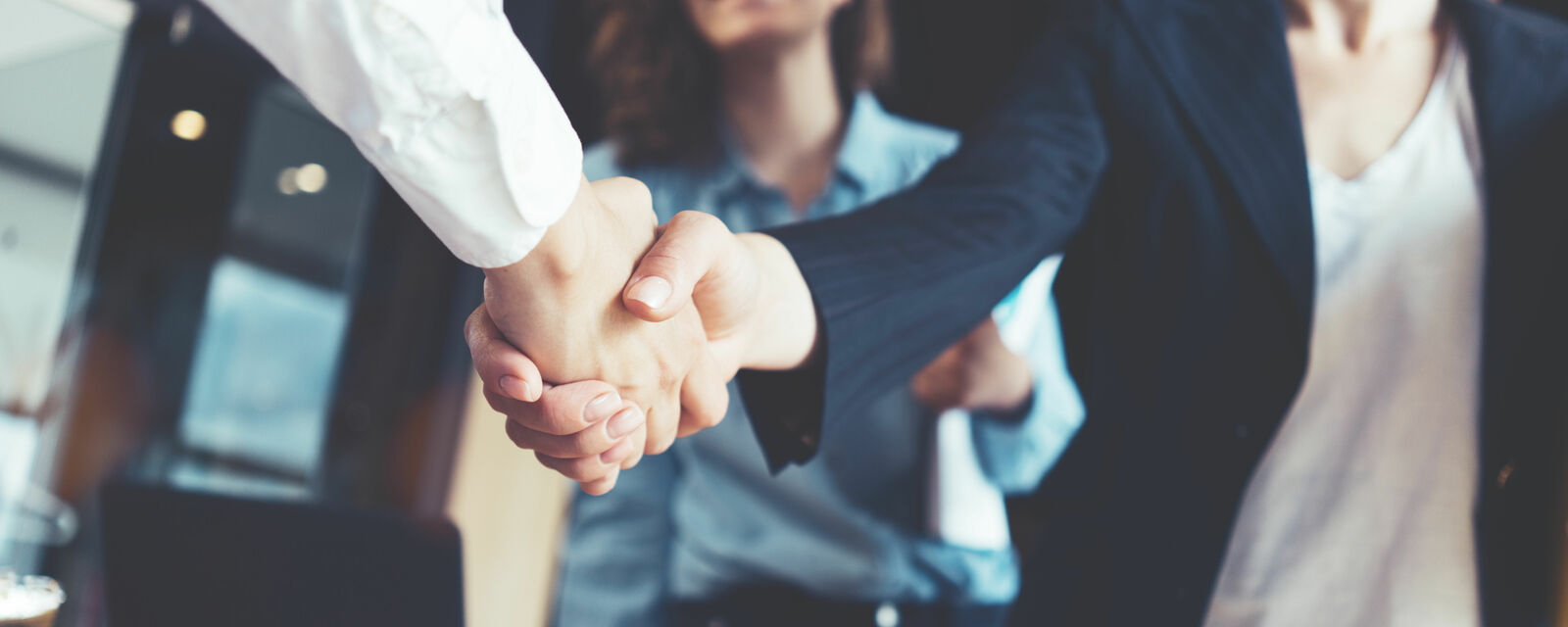 Close-up of a firm handshake between two people in a business setting. In the background, two other individuals in business attire are slightly out of focus. The scene conveys professionalism and collaboration.
