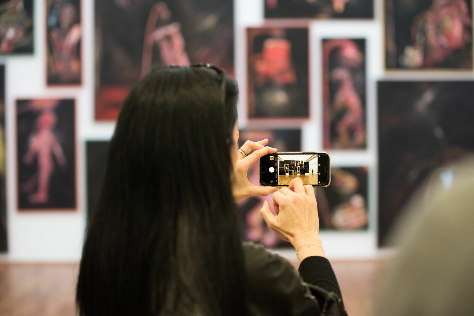 A person with long black hair takes a photo with a smartphone of an exhibition with artworks on the wall.