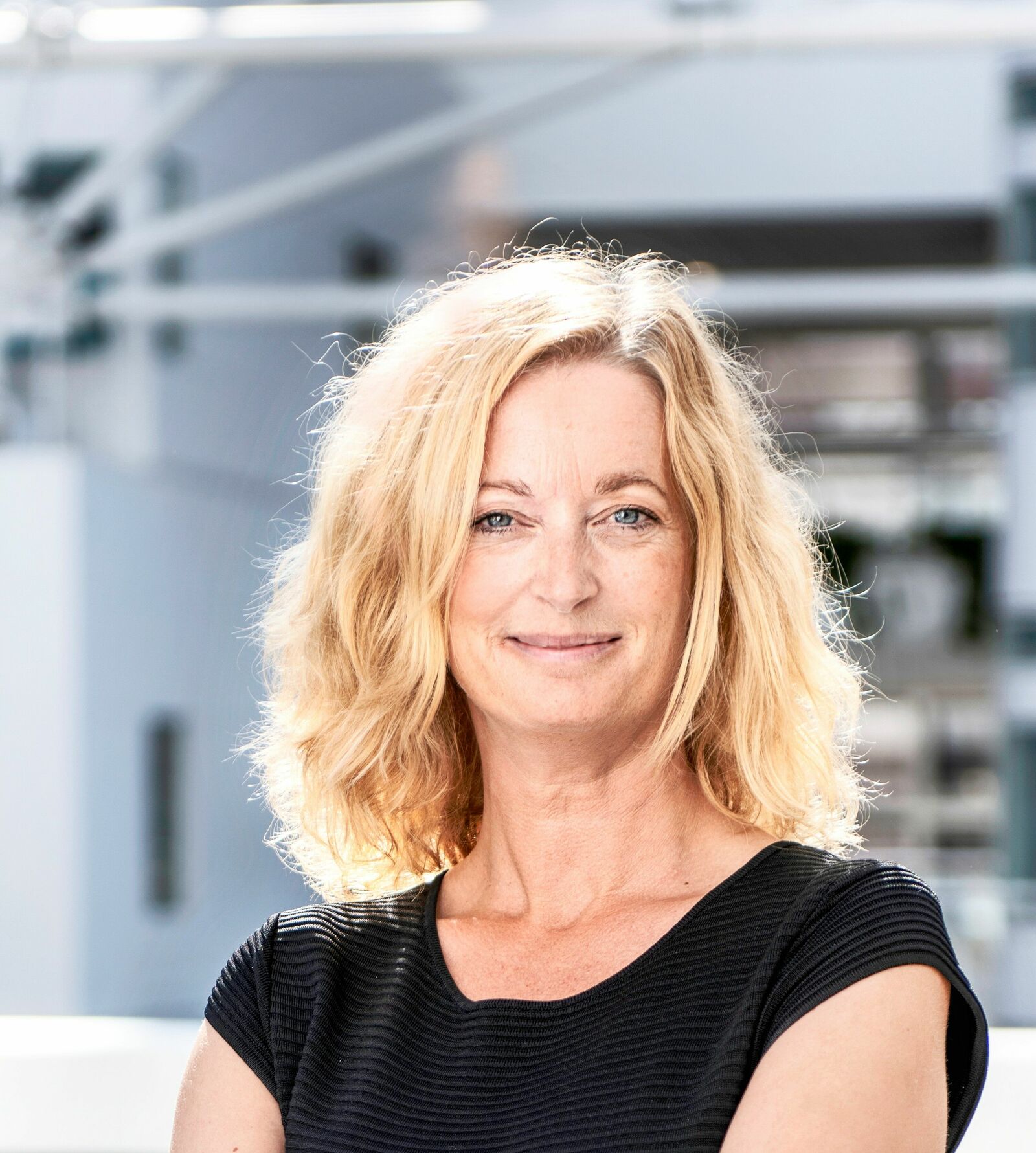 A woman with shoulder-length, wavy blonde hair smiles gently at the camera. She is wearing a black, textured top. The background is bright and modern, possibly an office environment with glass and metal structures.