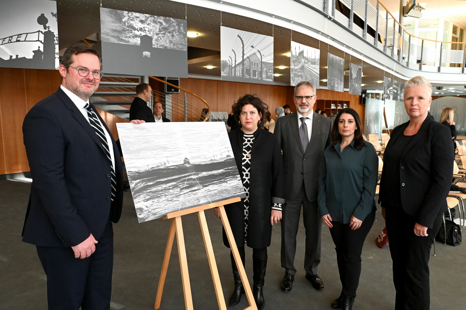 Five individuals stand in a modern exhibition space in front of a painting of a lighthouse by the sea displayed on an easel.