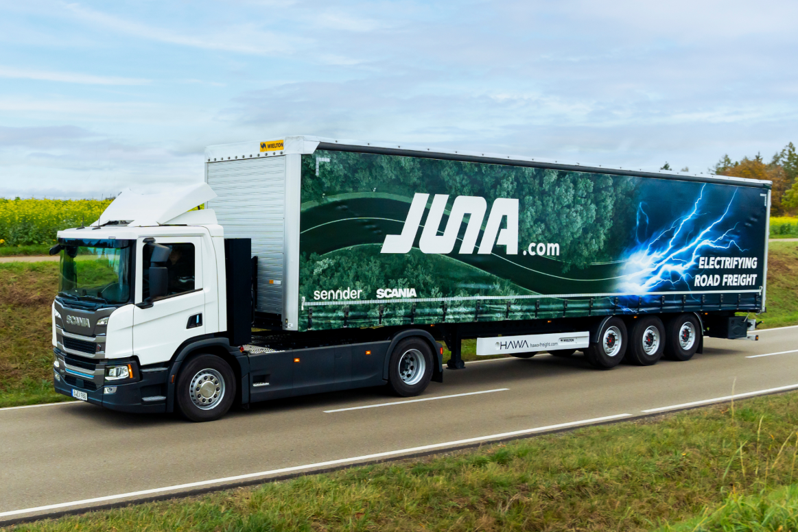 A white Scania truck with a large trailer drives on a country road. The trailer features the logo “JUNA.com” with a green forest design and blue lightning bolts, along with the slogan “ELECTRIFYING ROAD FREIGHT.”