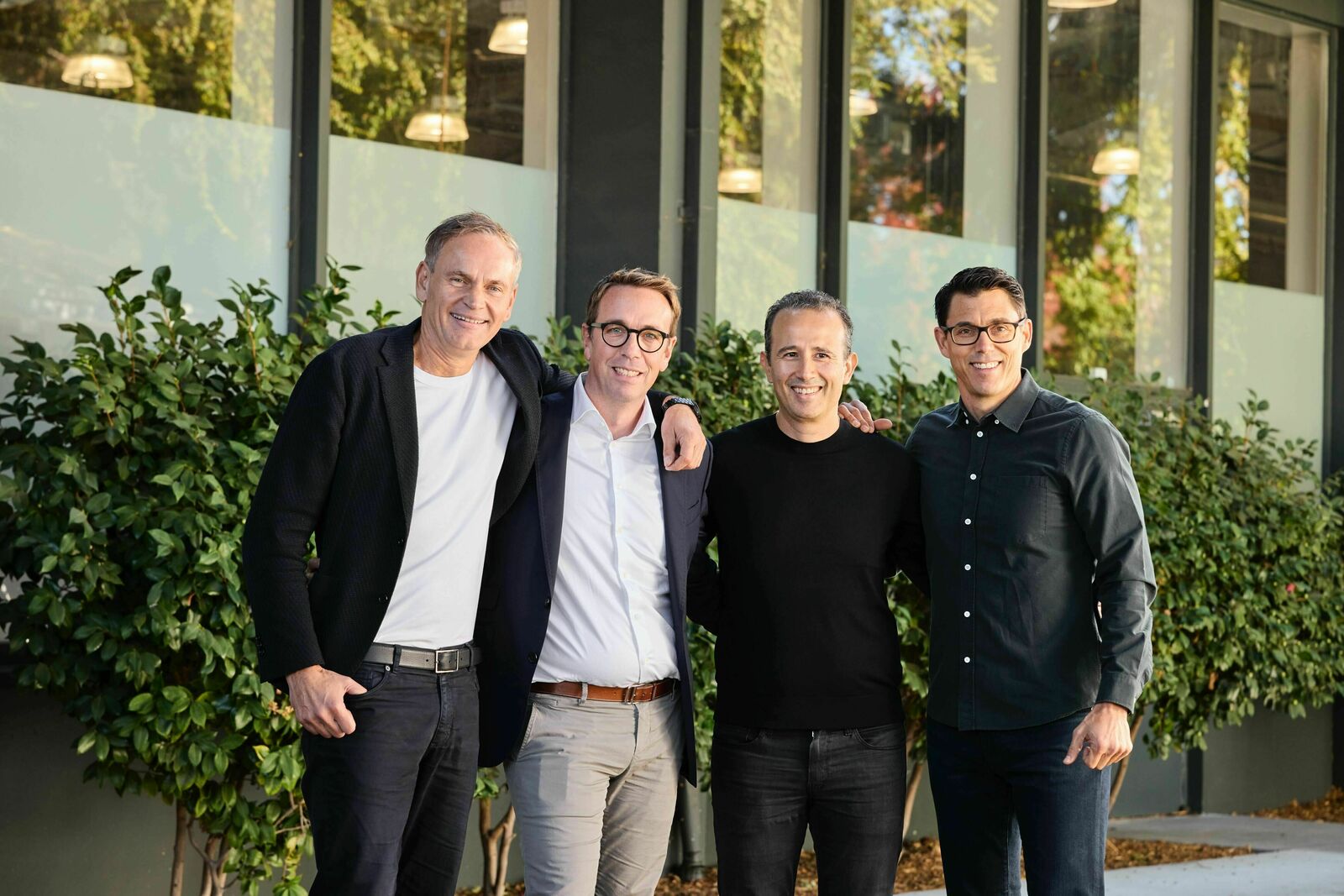 Four men stand close together, smiling at the camera. They are dressed in casual and business attire, standing in front of a modern building with large windows and green shrubs.