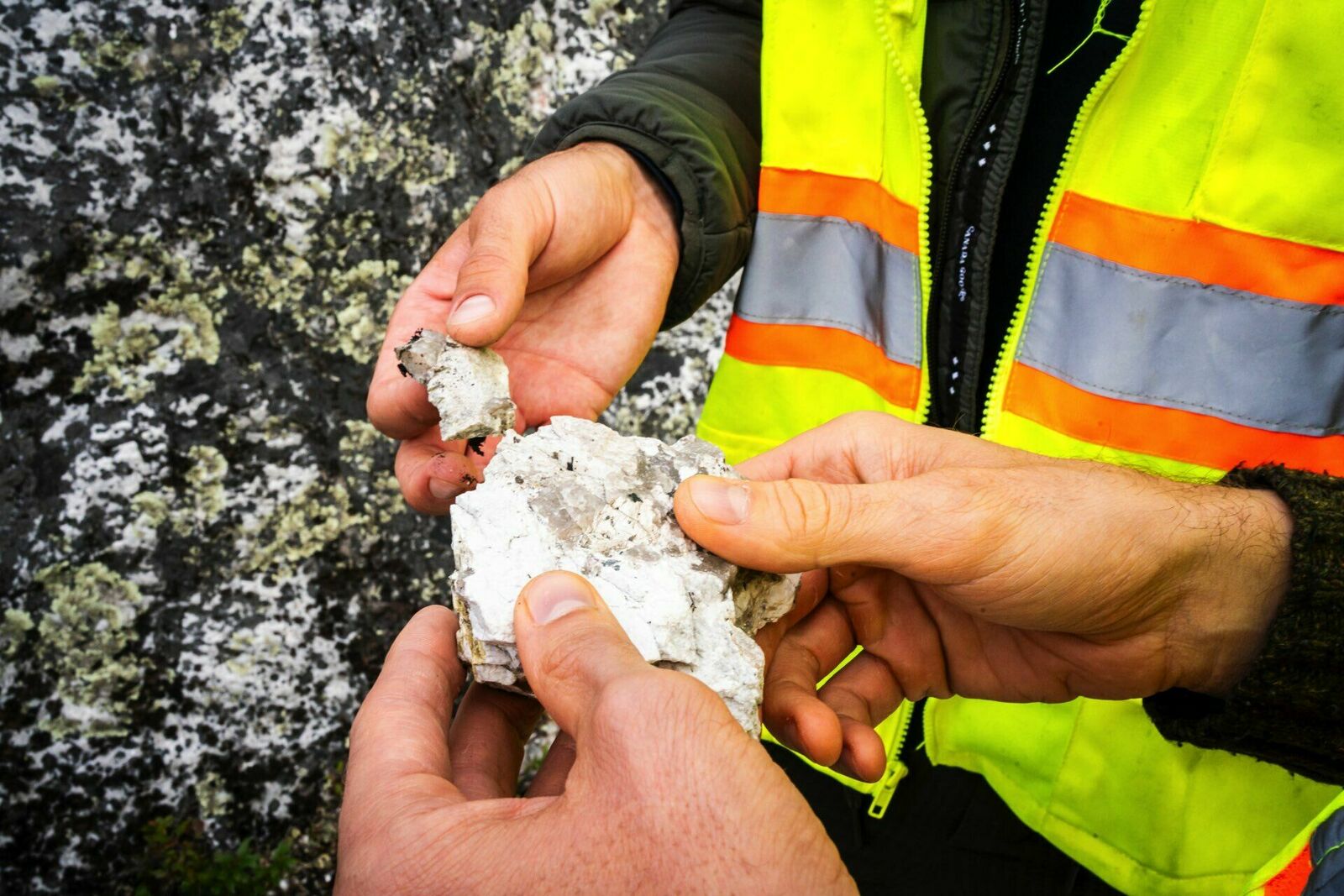 Close-up of two hands examining a rock fragment. One person is wearing a high-visibility vest with reflective stripes, suggesting work in construction, mining, or a similar industrial setting. The background features a rocky surface with lichen growth.