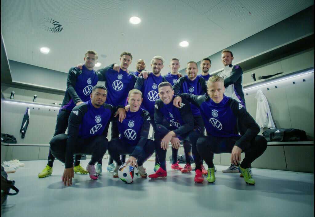 Eine Gruppe von elf Fußballspielern posiert lächelnd in einer Umkleidekabine für ein Teamfoto. Sie tragen einheitliche Trainingskleidung in dunklem Blau mit einem Logo und dem Volkswagen-Emblem auf der Vorderseite. Im Hintergrund sind offene Spinde, Jacken und Sporttaschen zu sehen. Ein Ball liegt zentral auf dem Boden vor den Spielern.