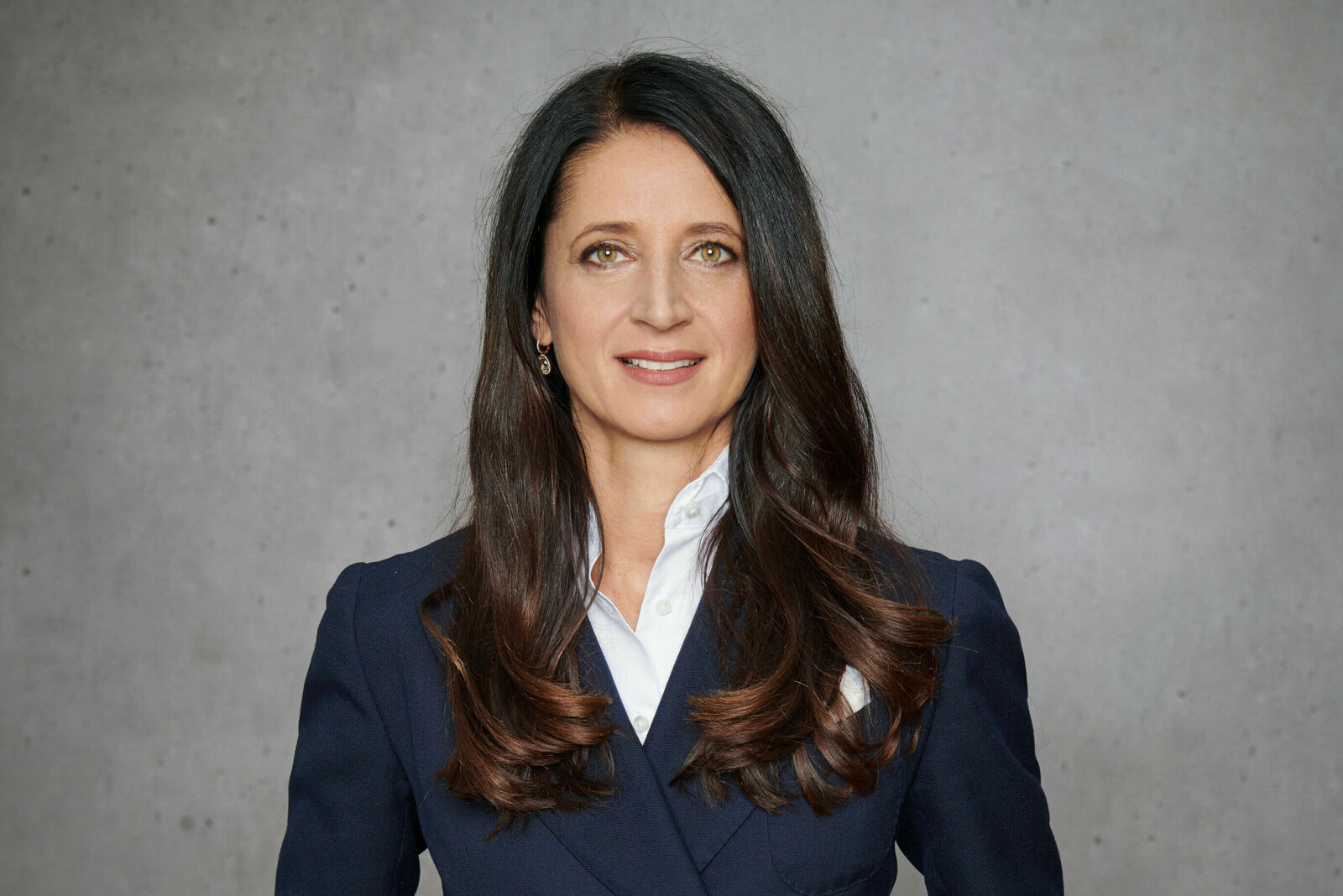 Das Bild zeigt eine professionelle Frau mit langen, dunklen Haaren, die in einem dunkelblauen Anzug mit weißem Hemd vor einer grauen Wand steht. Sie hat einen freundlichen Gesichtsausdruck.