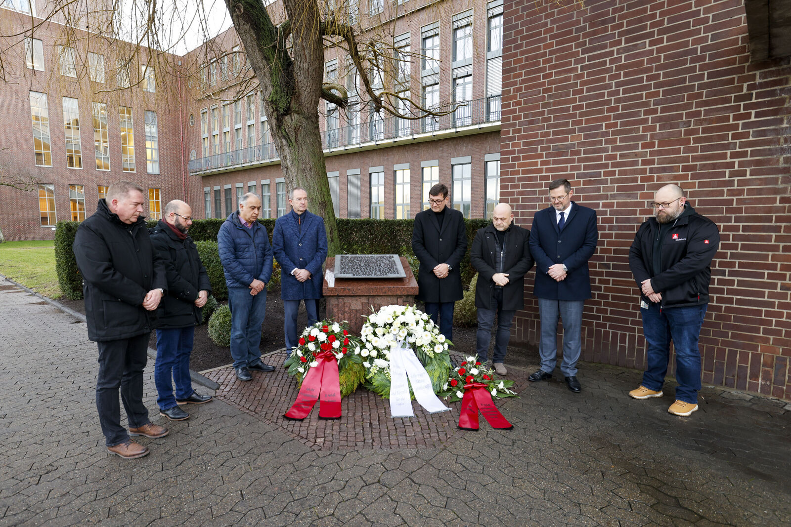 Eine Gedenkveranstaltung im Freien, bei der mehrere Personen in einer Reihe vor einem Denkmal stehen, umgeben von Blumenkränzen in Rot und Weiß. Die Teilnehmer senken ihre Köpfe in einer stillen Gedenkminute. Im Hintergrund sind Backsteingebäude und eine baumbestandene Umgebung zu sehen.