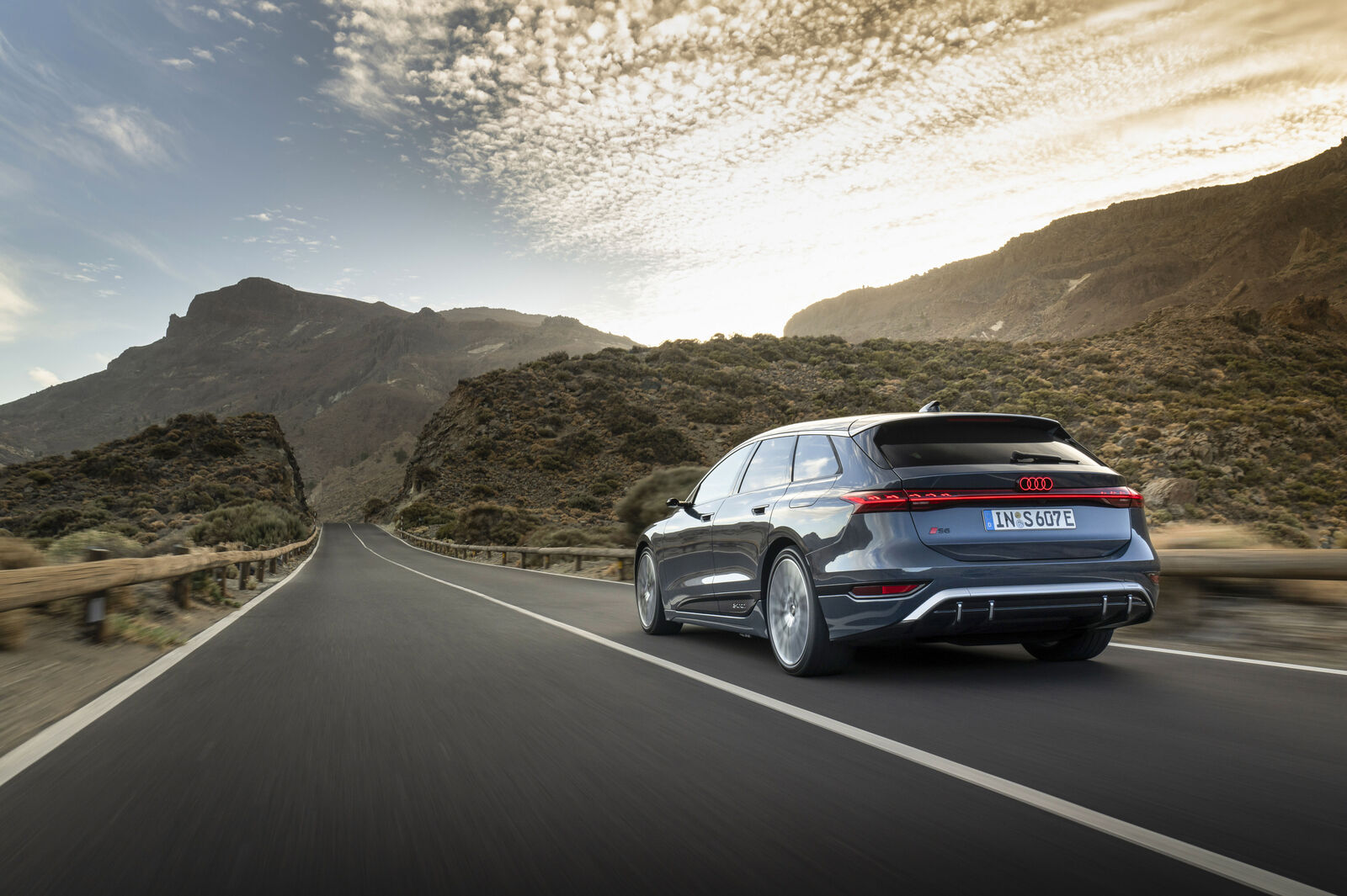 An Audi S6 Avant drives on a winding road through a mountainous landscape at sunrise or sunset.